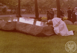 Jan Paweł II modli się przy grobie ks. J. Popiełuszki. Za nim grupa reporterów, fragment ogrodzenia kościoła pw. św. Stanisława Kostki. Za ogrodzeniem zebrany tłum. 13 VI 1987 r Sygnatura: IPNBU-7-14-11-12. Autor nieznany.